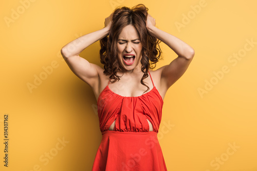 angry young woman screaming while standing with closed eyes and touching hair on yellow background