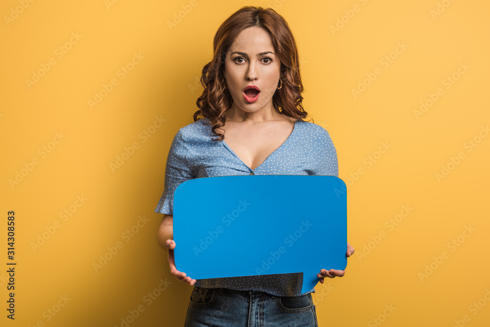 Fototapeta premium shocked girl looking at camera while holding speech bubble on yellow background