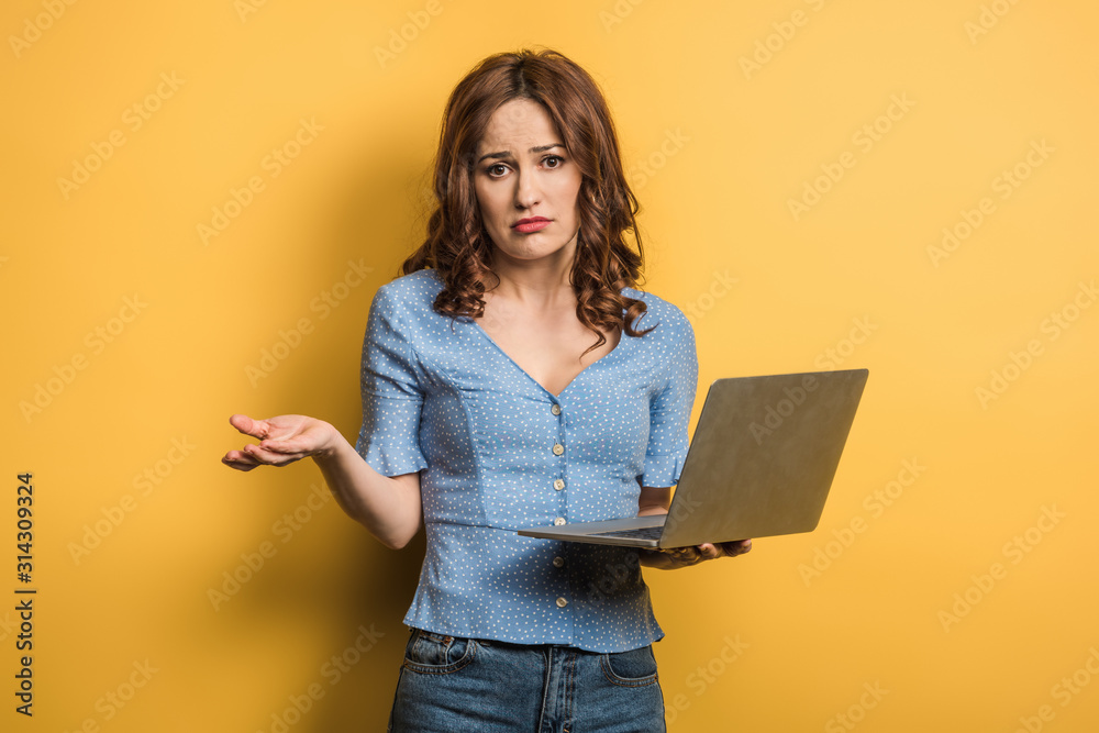 confused girl showing shrug gesture while holding laptop on yellow background confused girl showing shrug gesture while holding laptop on yellow background