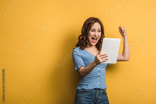excited woman showing winner gesture while holding digital tablet on yellow background photo