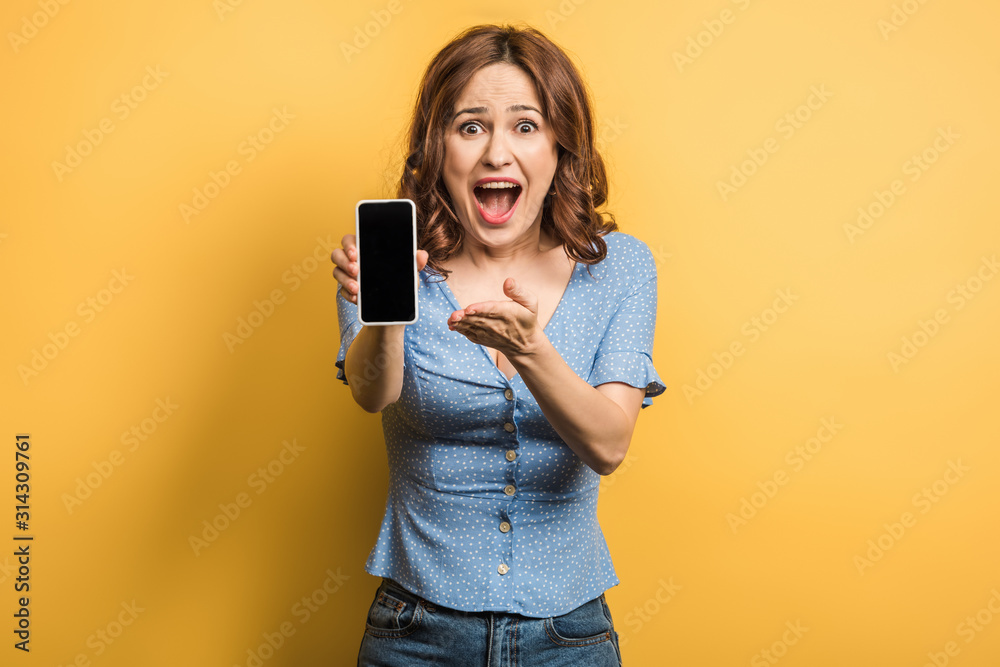 shocked woman pointing with finger at smartphone with blank screen on yellow background shocked woman pointing with finger at smartphone with blank screen on yellow background
