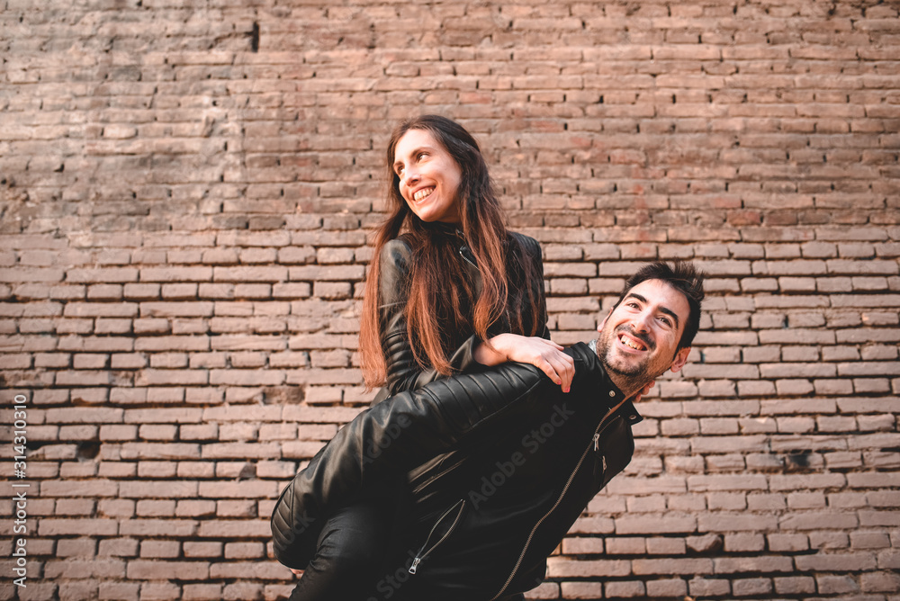 Φωτογραφία Young girl climb on the back of her boyfriend, piggyback, in front of a brick wall having fun and laughing loudly