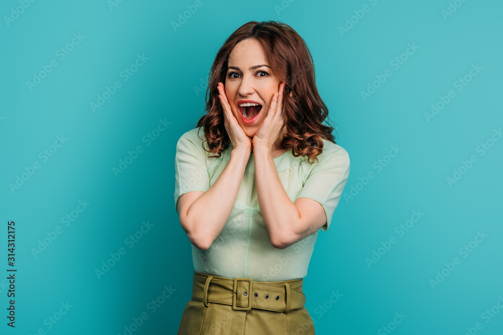 shocked woman touching face while standing with open mouth on blue background shocked woman touching face while standing with open mouth on blue background