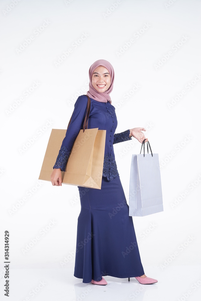 A beautiful and excited Muslim female model in a Asian traditional dress modern kebaya carrying shopping bags isolated on white background. Eidul fitri fashion and festive preparation shopping concept