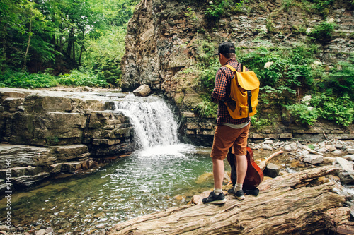 Traveler man with a backpack and a guitar stands on a trunk of a tree against a waterfall. Shoot from the back. Place for text or advertising