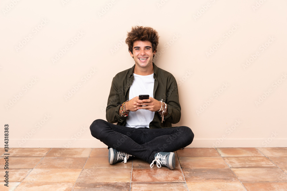 Young caucasian man sitting on the floor sending a message with the mobile