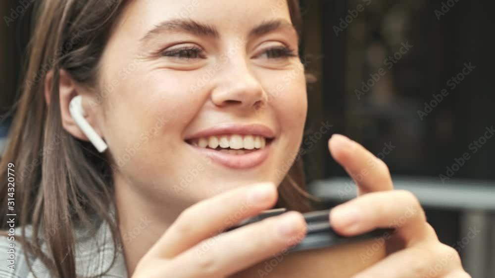 A pretty smiling young woman is talking with someone using wireless headphones while sitting at the caf� outdoors A pretty smiling young woman is talking with someone using wireless headphones while sitting at the caf� outdoors