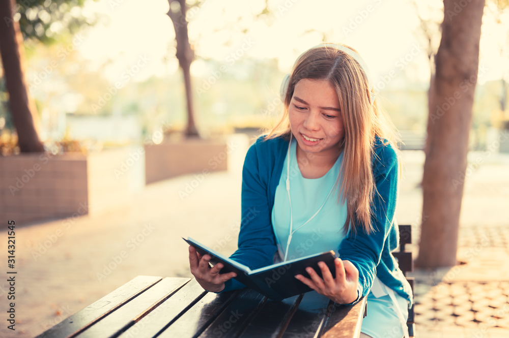 Obraz premium Smiling woman in eyeglasses reading book and listening to music with headphones in park outdoor against sunlight,City lifestyle concept.