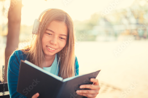 Smiling woman in eyeglasses reading book and listening to music with headphones in park outdoor against sunlight,City lifestyle concept.