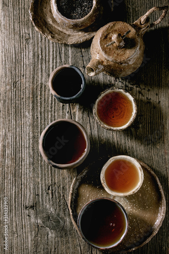 Hot black tea in different traditional wabi sabi style fireclay ceramic craft cups served with plate, teapot and bowl of dry tea leaves on old wooden table. Dark rustic atmosphere. Flat lay, space