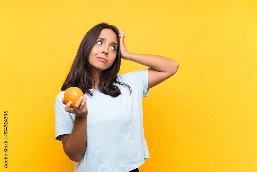 Young brunette woman holding an orange having doubts and with confuse face expression Young brunette woman holding an orange having doubts and with confuse face expression