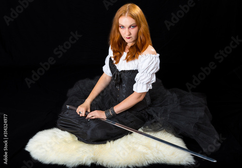 young woman wearing in white blouse, black long skirt and corset sitting with longsword on a sheepskin, on a dark background photo