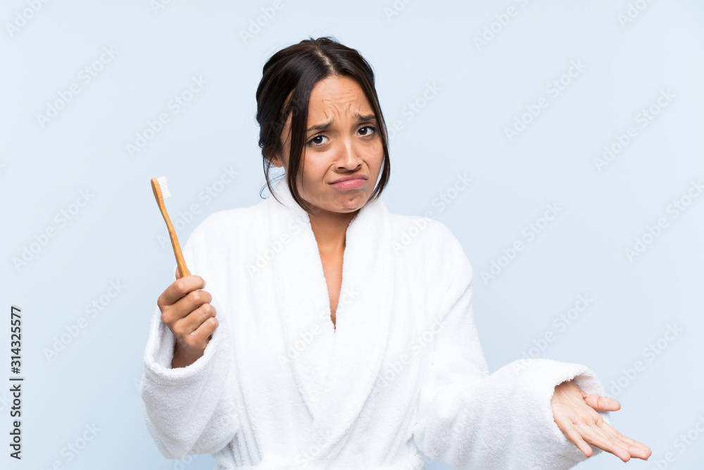 Young brunette woman in bathrobe brushing her teeth over isolated blue background making doubts gesture while lifting the shoulders Young brunette woman in bathrobe brushing her teeth over isolated blue background making doubts gesture while lifting the shoulders