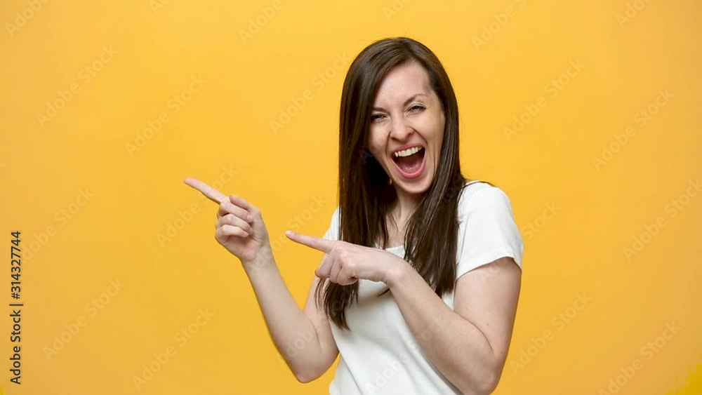Beautiful cute brunette young woman in white t-shirt showing thumbs up, pointing on copy space with charming smile isolated over yellow background in studio. People emotions lifestyle concept. Mock up