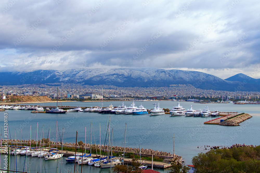 Aerial winter landscape view of harbor with moored luxury yacht and boats in marina of the city of Piraeus. Mountain rang in snow in the background. Attica, Greece