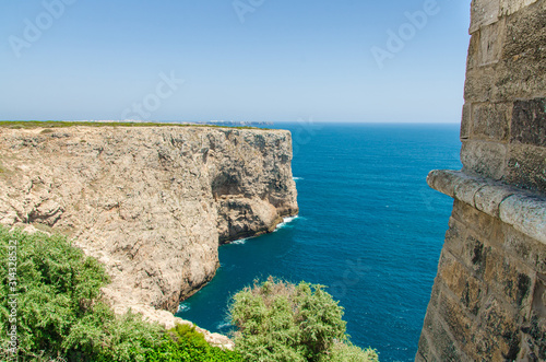 Portugal, Algarve, view of cliffs of Moher and Atlantic Ocean,  Cape St. Vincent on a sunny day with The blue Atlantic  in the background, stone wall in the foreground and green bushes in stones