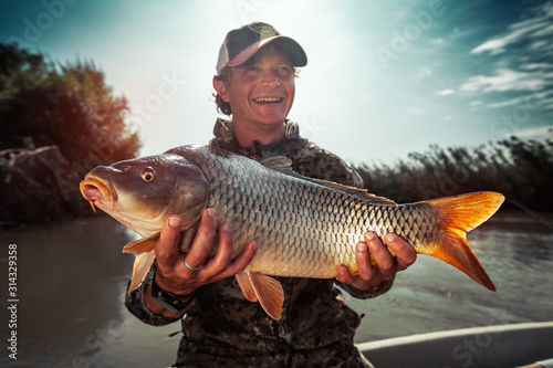 Fototapete Happy young fisherman holds the big Carp fish (Cyprinus carpio) and smiles