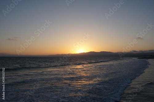 Sunsetting behind the mountains just off the beach of Santa Monica, California with a surfer and paraglider enjoying the views