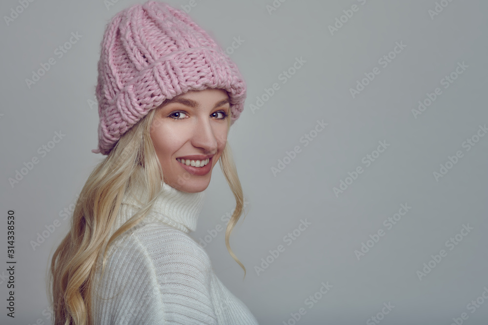 Happy young woman with long blond hair in pink knitted winter hat looking at the camera with a grey background Happy young woman with long blond hair in pink knitted winter hat looking at the camera with a grey background