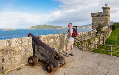 A hiker looks at the Atlantic from a castle in Galicia in northern Spain. There is a historic cannon in the foreground and a watchtower in the background. Sunshine and blue sky. photo