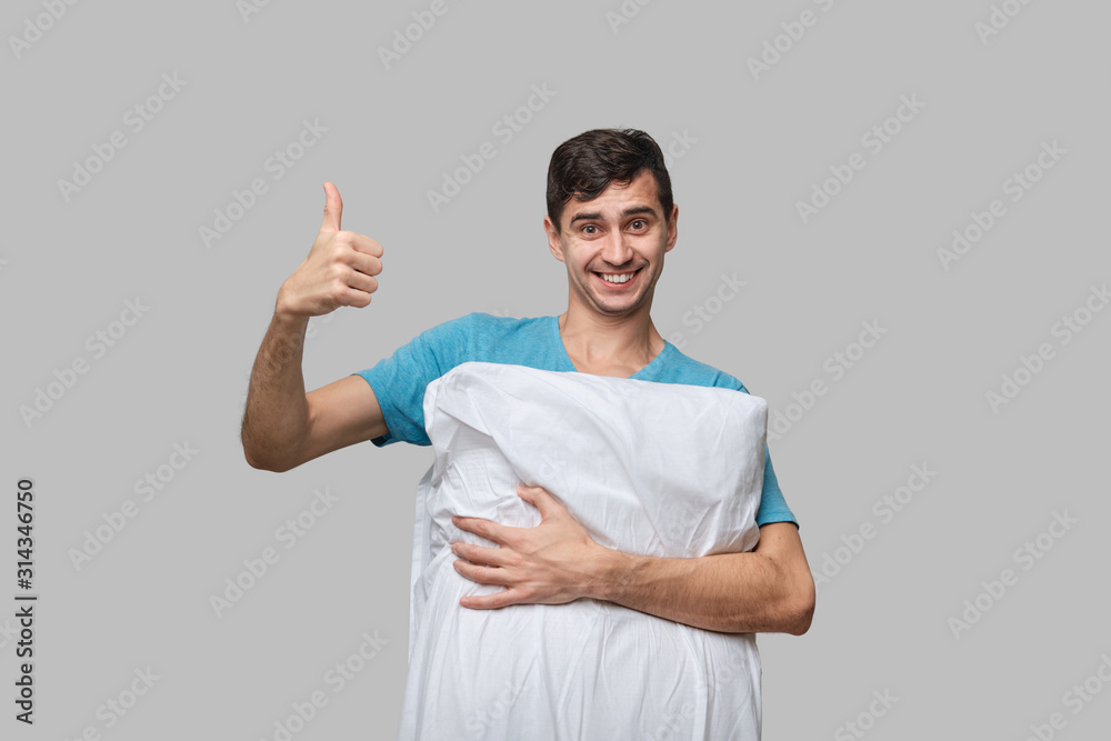Young brunet man shwoing thumb up holding white pillow isolated over grey background