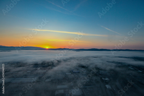 Aerial view of a foggy sunrise over a residential and industrial area of a city. Fog layered over houses below early morning sunrise.