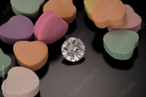A solitaire, faceted, round diamond is surrounded by candy hearts and sits on a reflective black background.