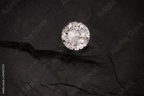 A solitaire, faceted, clear, round diamond sits on a dark rough slate background.