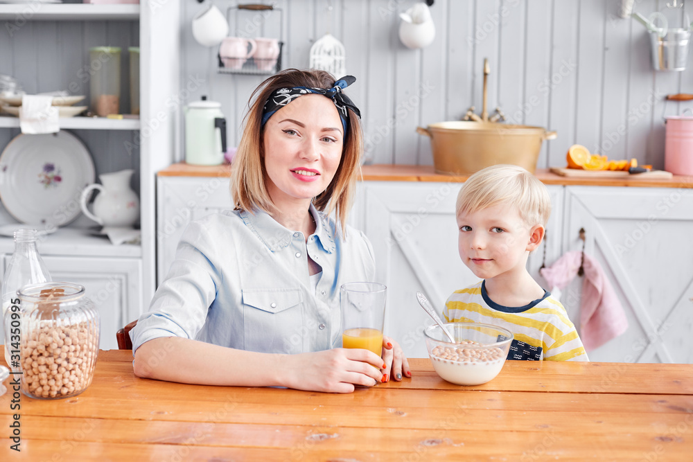 Mother and son are smiling while having a breakfast in kitchen. Bright morning in the kitchen. Healthy Breakfast cereals and fresh fruit. Mother and son are smiling while having a breakfast in kitchen. Bright morning in the kitchen. Healthy Breakfast cereals and fresh fruit.