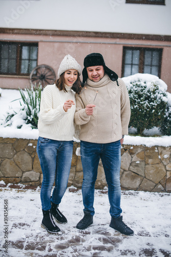 Beautiful young girl and guy in warm white sweaters having fun in the snowy yard of the house. It's Christmas time, the snow is falling.