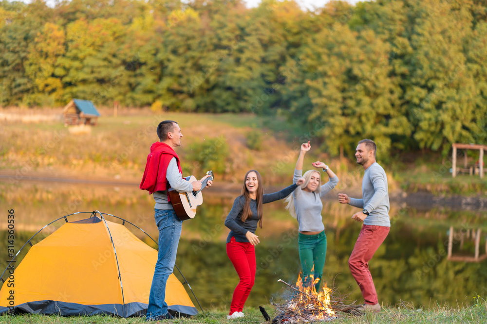 Group of happy friends with guitar, having fun outdoor, dancing and jumping near the lake in the park background the Beautiful sky. Camping fun