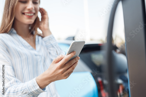 Transportation. Young woman on electric car having stop at charging station using app on smartphone close-up blurred background cheerful while getting car fully charged