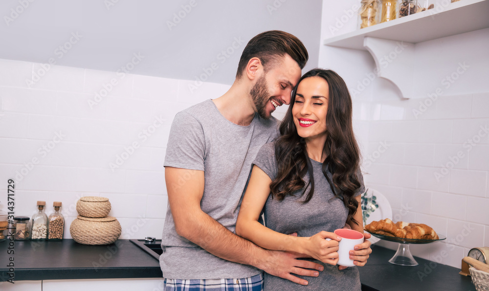 Kitchen romance. Close-up photo of a wonderful woman, who is holding a cup of coffee, and a handsome man, who is hugging her from the back in the kitchen.