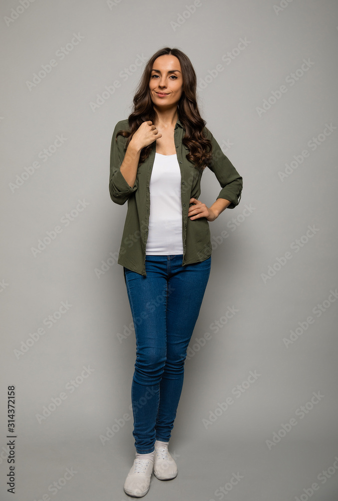 Perfect hair. Full-length photo of a dark-haired woman in a khaki shirt and blue jeans, who is posing in front with her left hand on her hip, looking in the camera and smiling. Perfect hair. Full-length photo of a dark-haired woman in a khaki shirt and blue jeans, who is posing in front with her left hand on her hip, looking in the camera and smiling.