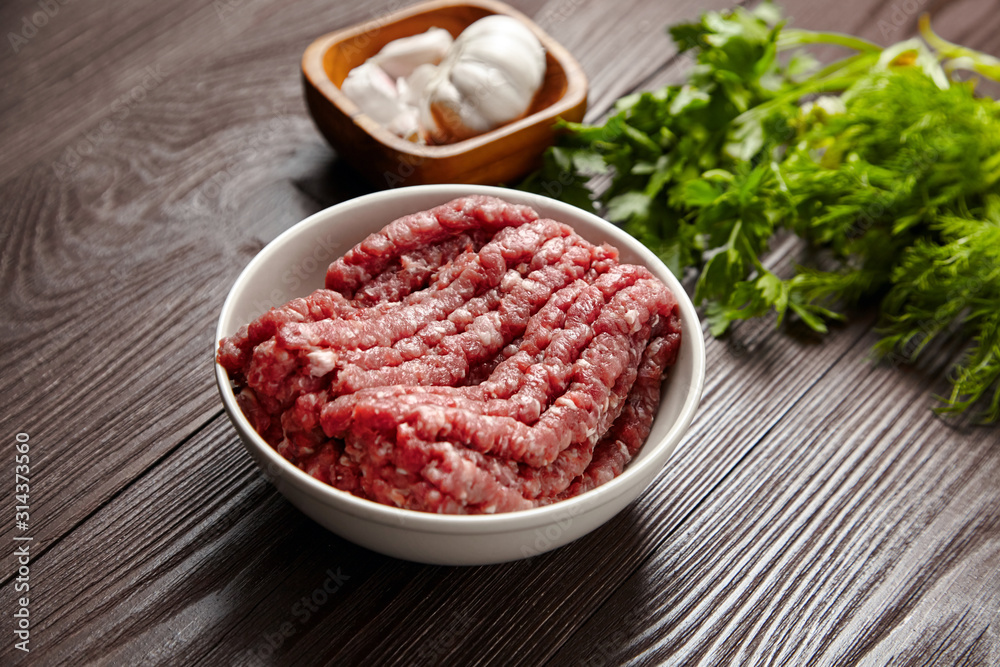 Bowl with raw ground beef and condiments on wooden table, closeup. Cooking minced meat with fresh seasonings. White garlic heads and cloves, green dill and parsley leaves Bowl with raw ground beef and condiments on wooden table, closeup. Cooking minced meat with fresh seasonings. White garlic heads and cloves, green dill and parsley leaves