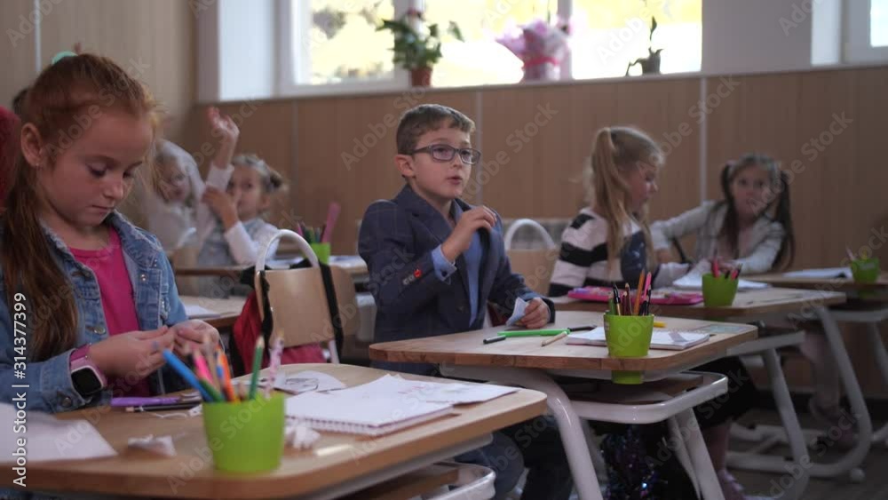 Cute elementary age boy in glasses raising hand and answering teacher's question during lesson at school. Smart primary school students wanting good grades and hurrying to raise hands in classroom Cute elementary age boy in glasses raising hand and answering teacher's question during lesson at school. Smart primary school students wanting good grades and hurrying to raise hands in classroom