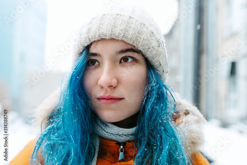 Young asian female with dyed blue hair in a white knitted hat standing in a snowfall, selective focus