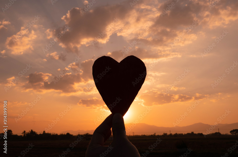 Fototapeta premium Silhouette of the heart shaped cardboard in hand and the sun shining through the heart on sunset background. Concepts of love and valentines day.