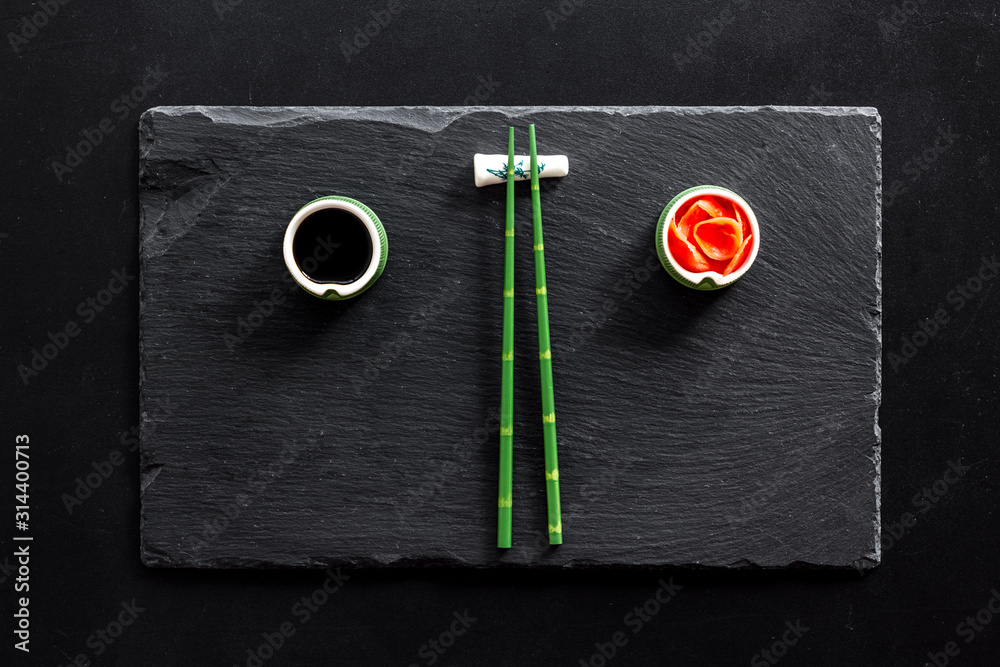 Table served for eating sushi. Chopsticks, small bowls with ginger and sause, mat on black background top-down