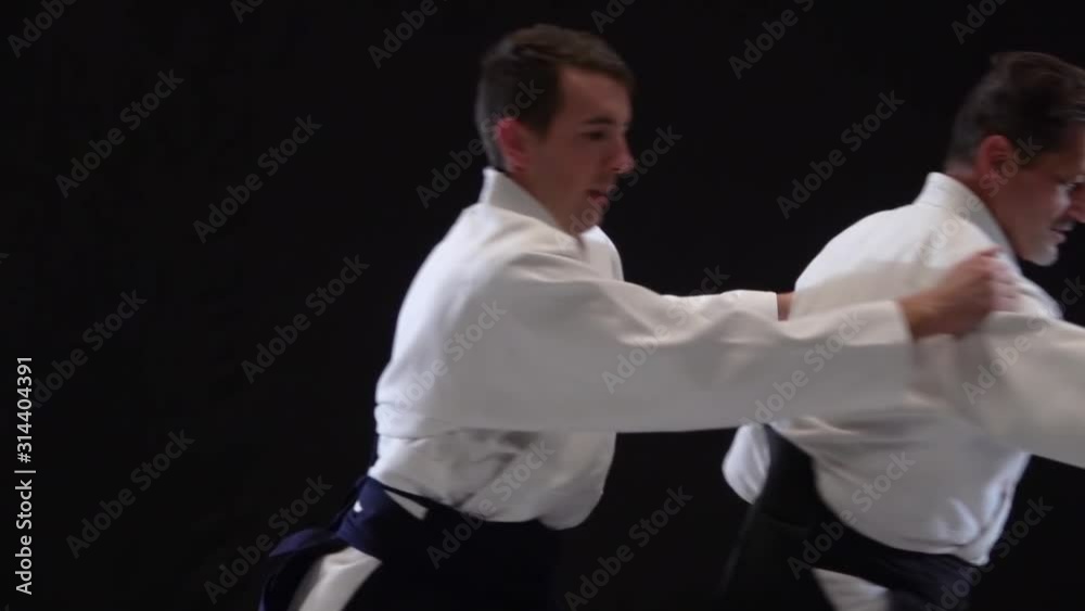 Two Masters participants of the training in special clothes of aikido hakama work out the methods of single combat on spotlights background. Slow motion. Close up. Two Masters participants of the training in special clothes of aikido hakama work out the methods of single combat on spotlights background. Slow motion. Close up.