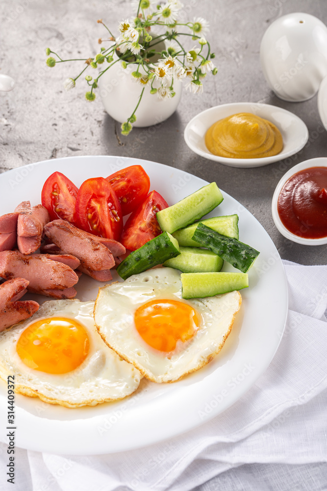 Homemade delicious breakfast with sunny side up fried egg, sausage, vegetable, jam, black coffee on a stone table. Breakfast served with flowers and white dishes Homemade delicious breakfast with sunny side up fried egg, sausage, vegetable, jam, black coffee on a stone table. Breakfast served with flowers and white dishes