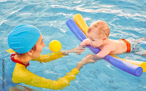 Young mother aged 30 years and baby son of 5 months are in pool. Smiling woman with beautiful snow-white smile is dressed in fashionable swimming suit swimsuit and cap. Girl teaches boy to swim.