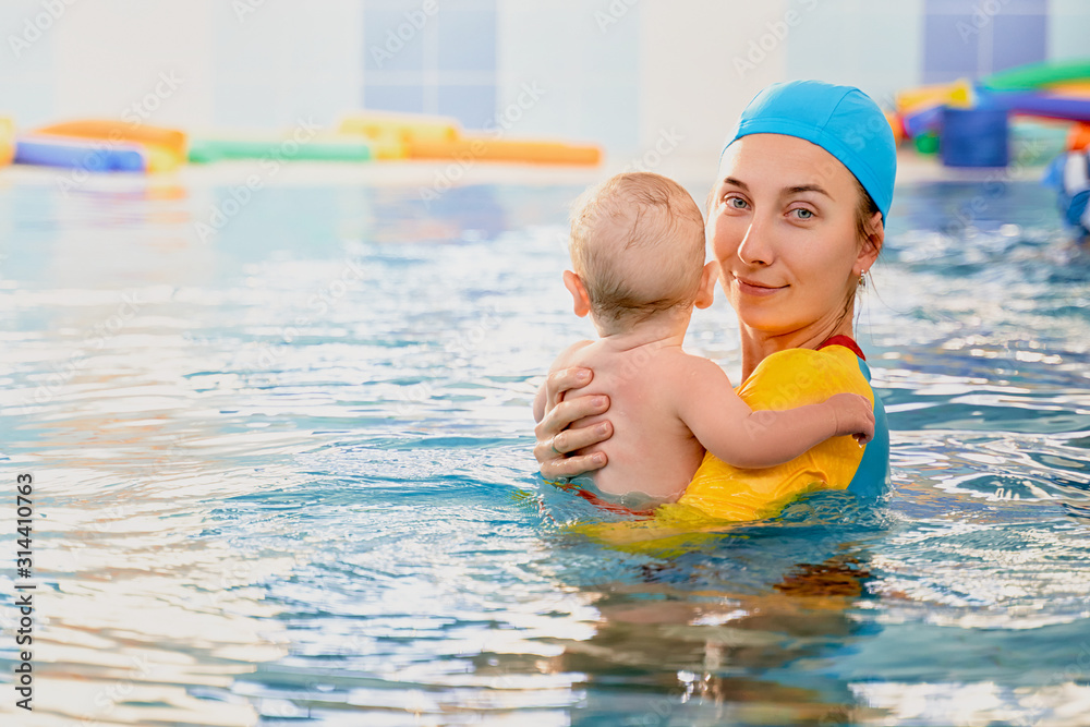 Young mother aged 30 years and baby son of 5 months are in pool. Smiling woman with beautiful snow-white smile is dressed in fashionable swimming suit swimsuit and cap. Free space for advertising