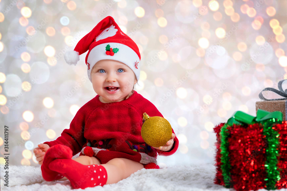 Portrait of a cute, blue eyed baby girl with a santa hat holding a golden christmas ornament in front of blurred, festive lights Portrait of a cute, blue eyed baby girl with a santa hat holding a golden christmas ornament in front of blurred, festive lights