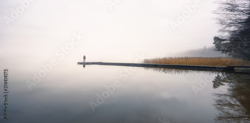 Man standing alone on edge of pier and staring at lake. Mist over water. Foggy air. Early chilly morning in autumn. Beautiful freedom moment and peaceful atmosphere in nature. Back view.