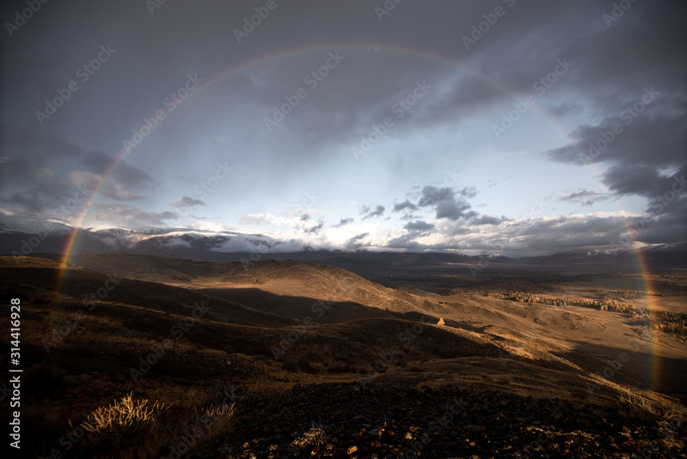 gloomy landscape of high gray mountains at sunset under the clouds with glimpses of bright sunlight and a full vibrant color rainbow