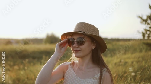 Portrait of pretty caucasian girl in hat is putting on sunglasses and smiling. Rural landscape at sunset and stylish young woman model posing. Summer evening