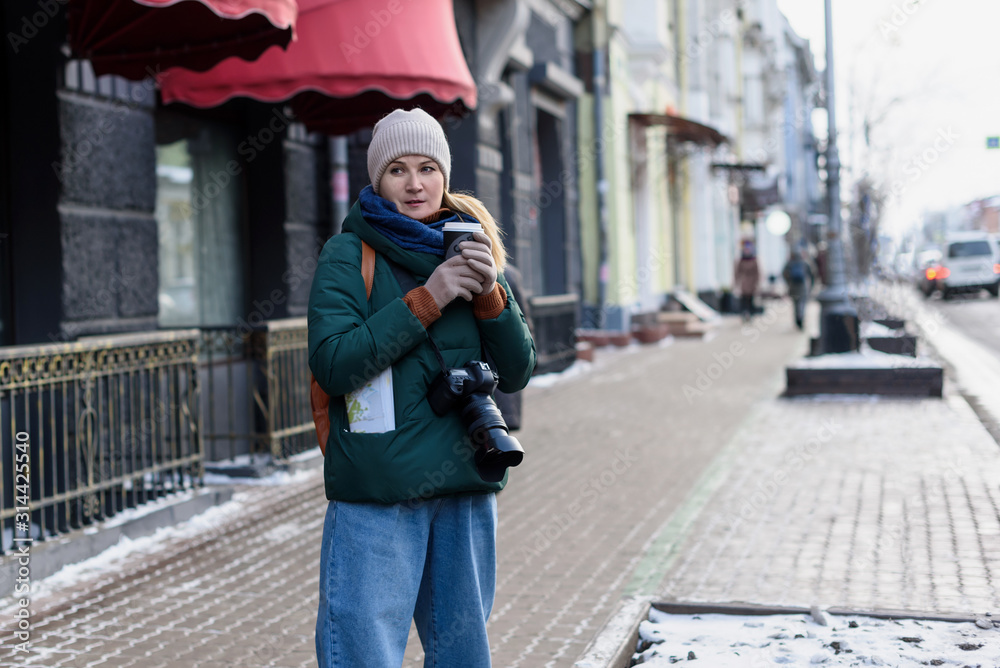 Girl in winter clothes with camera and map in the historical center of the city is holding a cup of coffee on the street