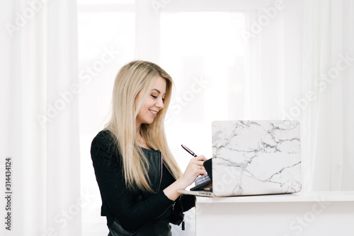 Beautiful smiling blonde girl with a notebook and pen in her hands, next to a laptop on a light background, office employee