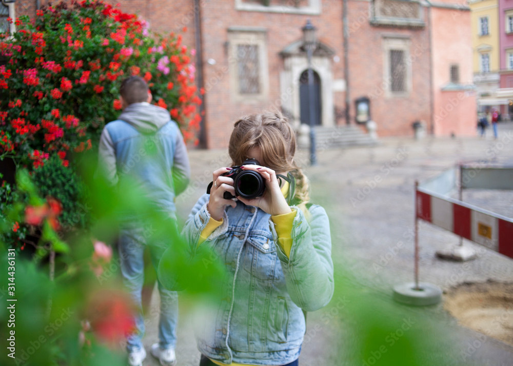  A girl in a denim jacket takes pictures, and a guy sniffs flowers in the background.